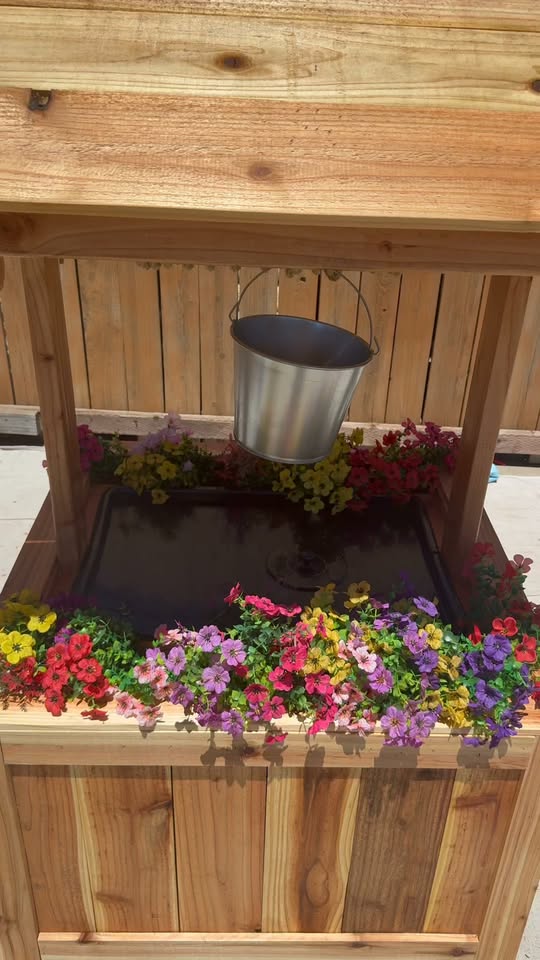 Close view of a cedar wishing well planter box with a galvanized bucket hanging over a black liner and flowers around the rim.